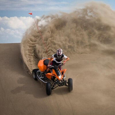 A person wearing a helmet and protective gear rides an orange ATV at the Killpecker Sand Dunes in 怀俄明. The ATV tires kick up sand and create a wavy wall behind it.