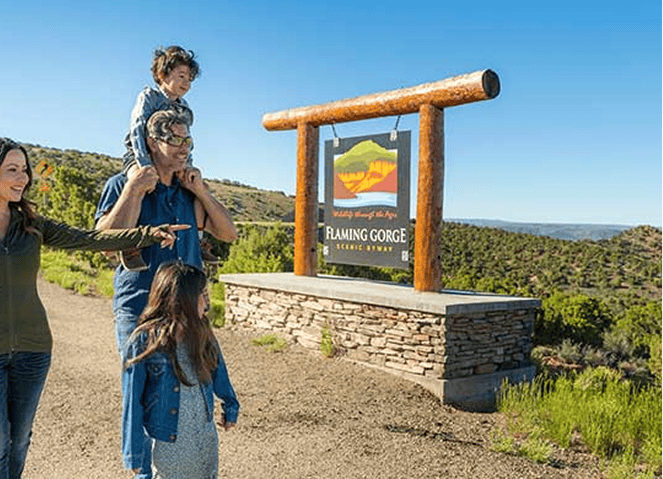 A family stands near a large sign for Flaming Gorge Scenic Byway, with hills and blue sky in the background.