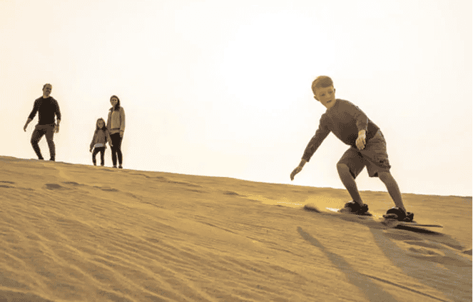 A boy sandboarding down a dune with three people standing at the top in the background under a bright sky.