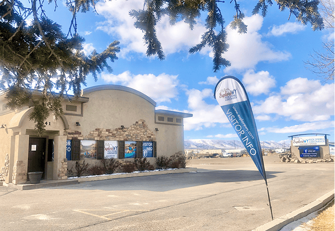 A beige building with "Visitor Info" signs and a blue flag in the parking lot; mountains and clouds visible in the background.