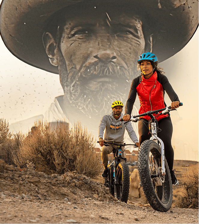 Two people ride mountain bikes on a dirt trail in front of a large black-and-white mural of a man's face wearing a hat.