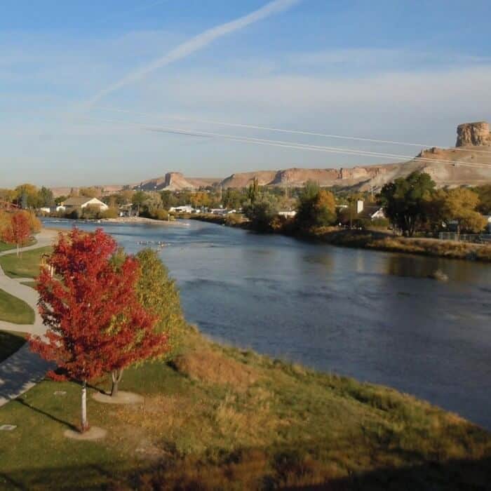 A wide river winds through a small town with autumn trees; rocky plateaus and a clear blue sky are visible in the background.