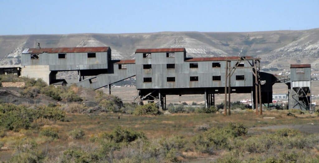 Abandoned industrial building with rusted metal siding and multiple levels, set in a dry, brushy landscape with hills in the background.