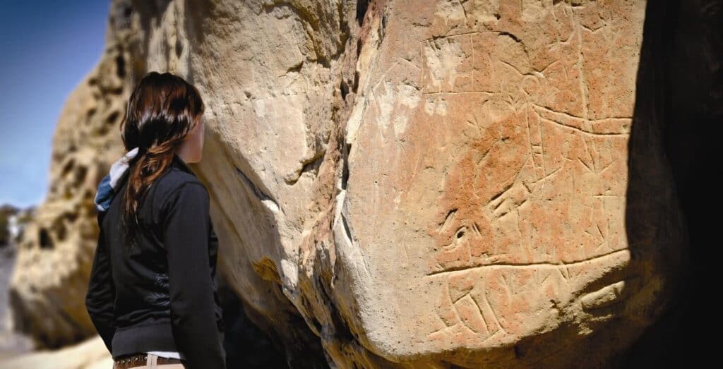 A person looks at ancient carvings on a large rock outdoors under natural light.