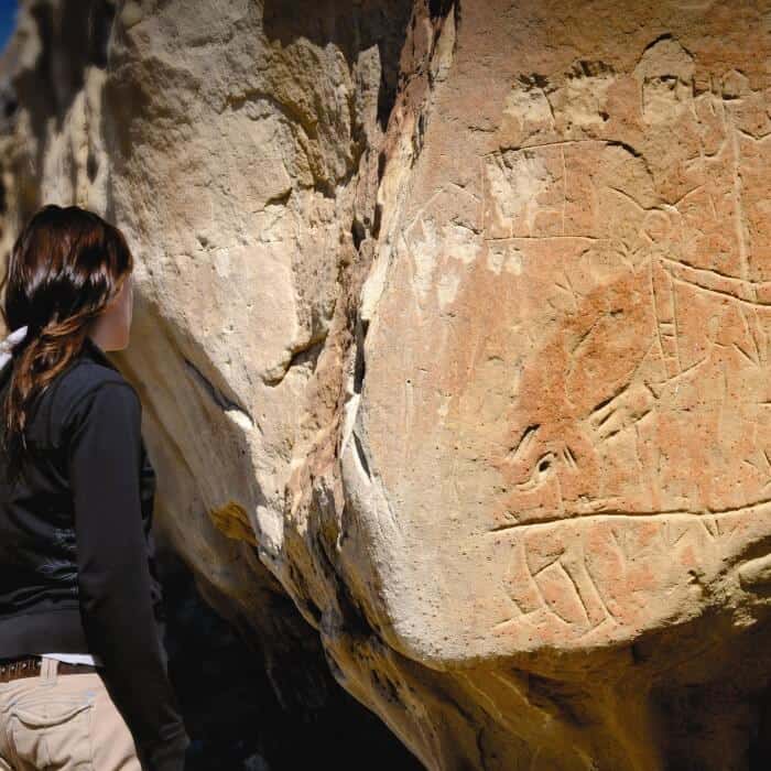 A person looks at ancient petroglyphs carved into a large rock surface outdoors.