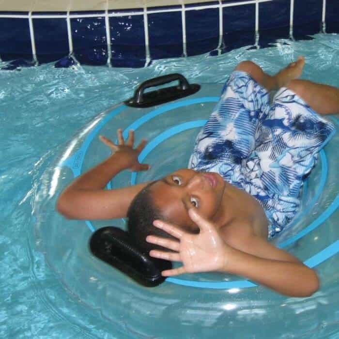 A boy in blue patterned swim trunks floats on a clear inner tube in a swimming pool, raising his hands and looking at the camera.