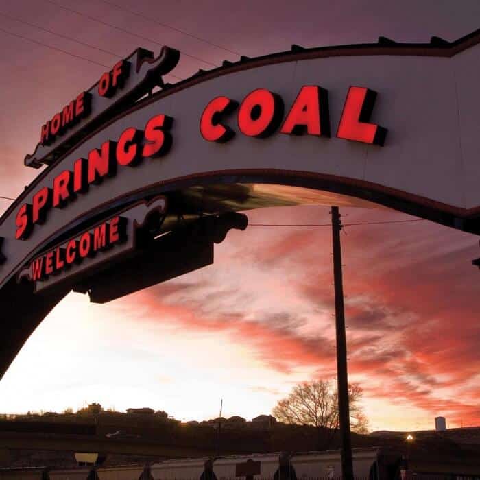 An illuminated sign reads "Home of Springs Coal Welcome" against a sunset sky with some silhouetted trees and buildings in the background.