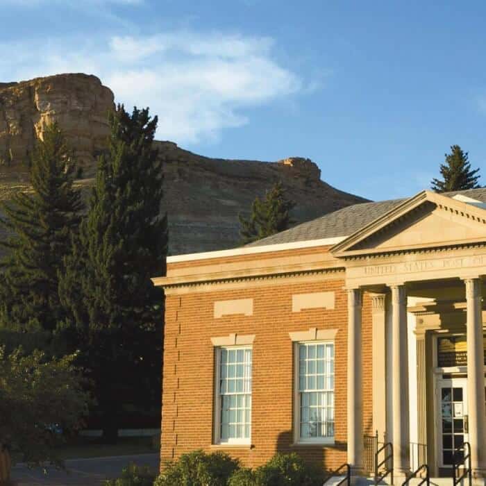 A brick post office building with columns stands in front of tall pine trees and a rocky hill under a blue sky.