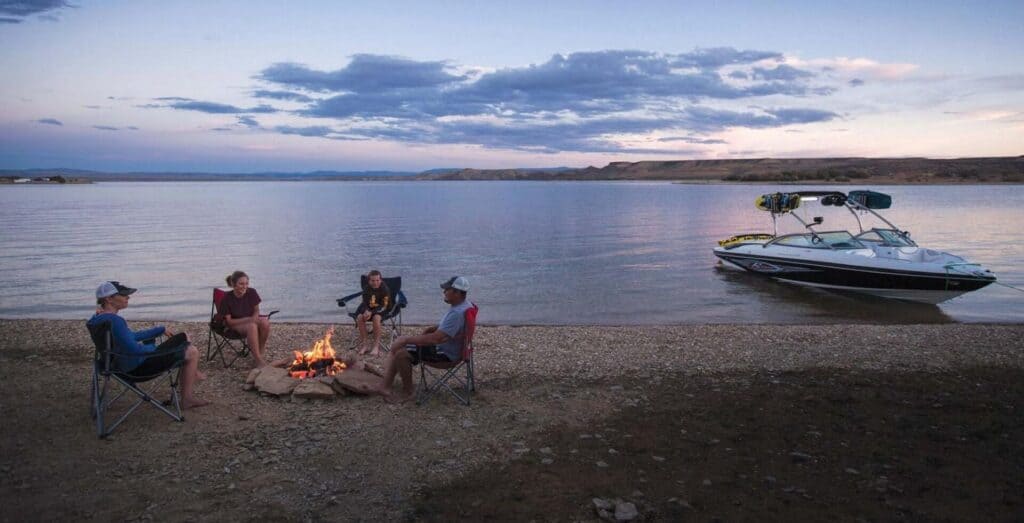 Four people sit around a campfire on a lakeshore at dusk, with a motorboat anchored nearby on the water.