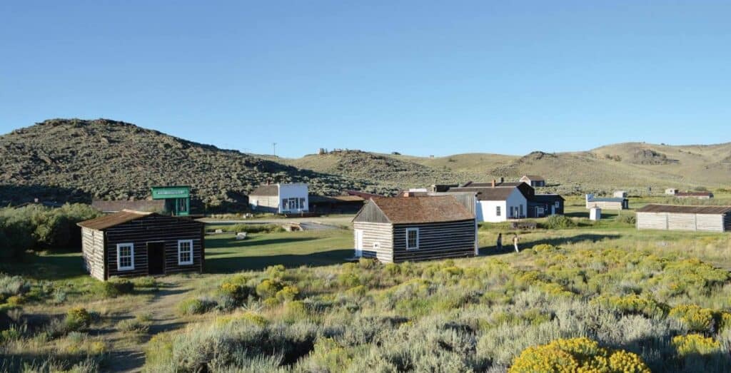 A cluster of small wooden buildings stands in a grassy, hilly landscape under a clear blue sky.