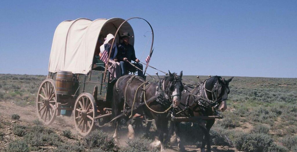 A covered wagon pulled by two horses travels on a dirt trail through a grassy landscape. Two people, wearing hats, ride inside. American flags are attached to the wagon.