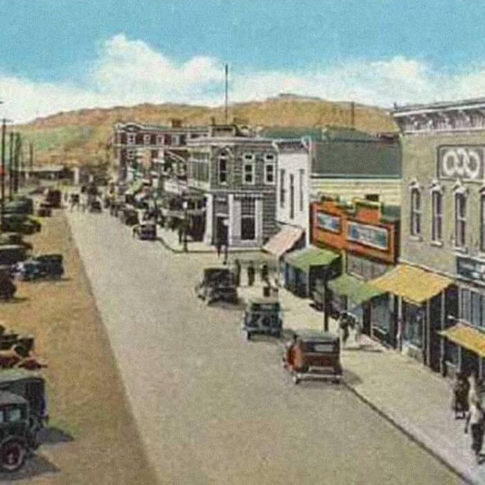 Colorized vintage photo of a small town main street with early automobiles, storefronts, pedestrians, and a distant hill under a partly cloudy sky.
