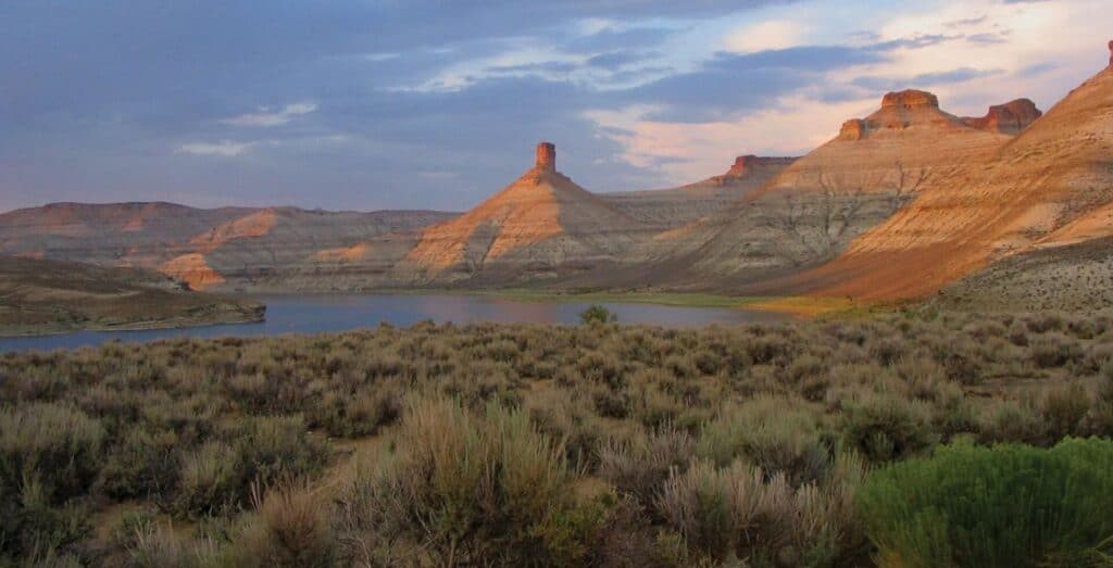 Desert landscape with sparse shrubs in foreground, a lake in the midground, and layered rocky mesas and buttes under a partly cloudy sky in the background.