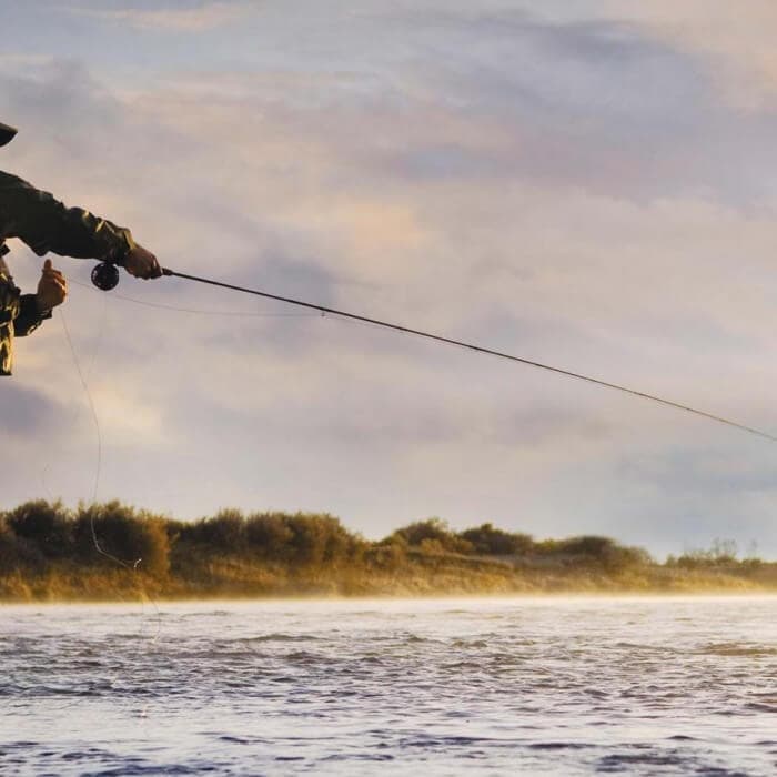 Person fishing in a river at sunrise or sunset, with the fishing rod extended over the water and a misty landscape in the background.