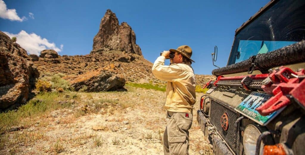 A person in outdoor gear uses binoculars near a rugged vehicle in a dry, rocky landscape with a tall rock formation under a clear blue sky.