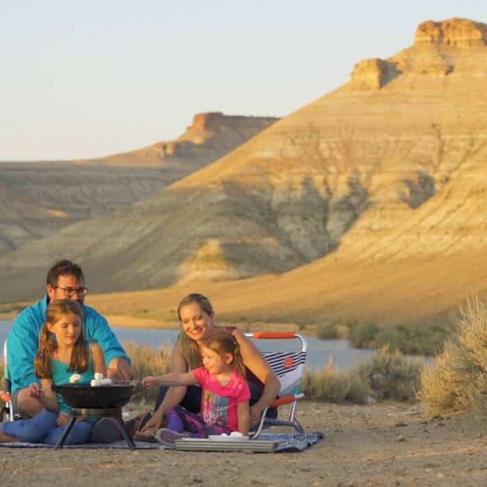 A family of four sits outdoors in a desert landscape, roasting marshmallows over a portable grill with rocky hills in the background.