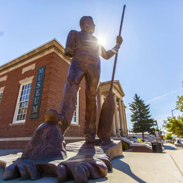 Bronze statue of a man holding an oar stands outside a brick building with a "Museum Free" sign, under a clear sky.