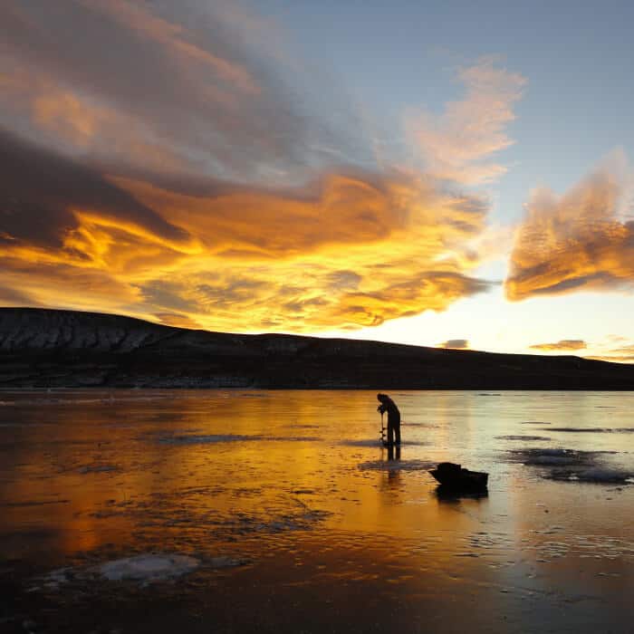 A person stands on a reflective frozen lake at sunset, with dramatic clouds and orange sky above dark hills.