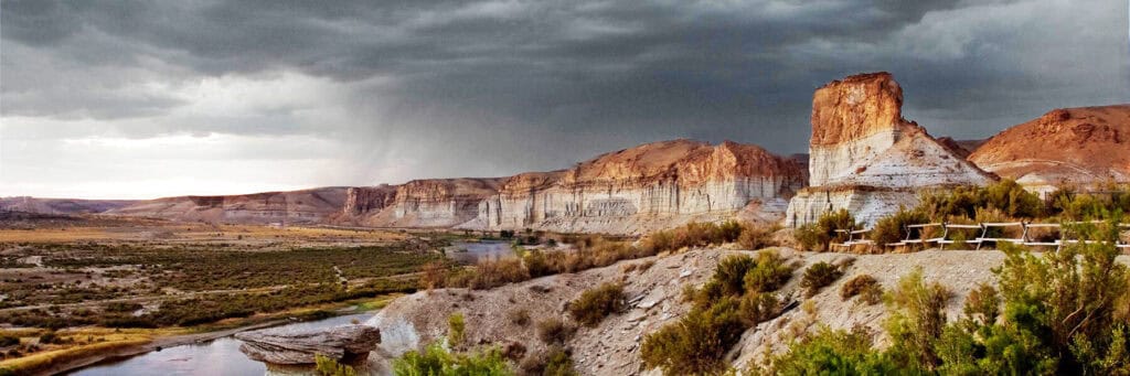 Rock formations and cliffs rise above a winding river under a cloudy sky, with sparse vegetation covering the rocky landscape.