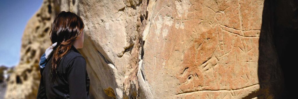 A person stands close to a large rock face featuring ancient carvings or petroglyphs under bright daylight.