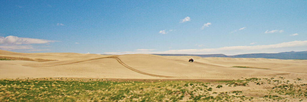 A lone off-road vehicle drives over sand dunes under a clear blue sky, with desert plants in the foreground and mountains in the distance.