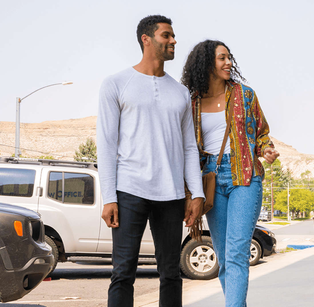 A man and woman walk together on a sidewalk near parked cars on a sunny day. Both are smiling and casually dressed.