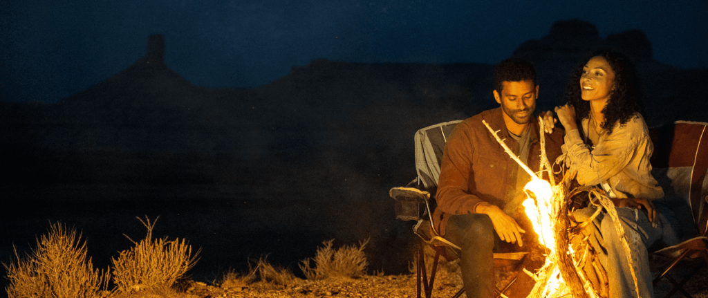Two people sit in camping chairs by a campfire at night in a desert landscape, with silhouettes of mesas visible in the background.