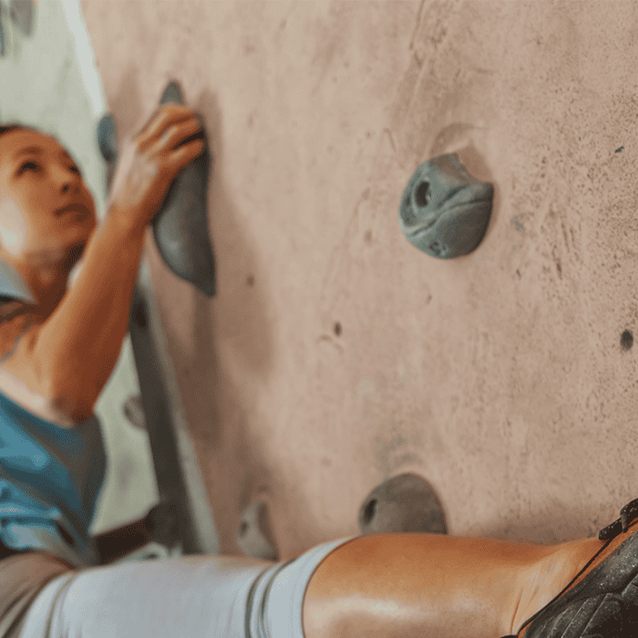 Person climbing an indoor rock wall, gripping a handhold with one hand and wearing climbing shoes, with a focused expression.