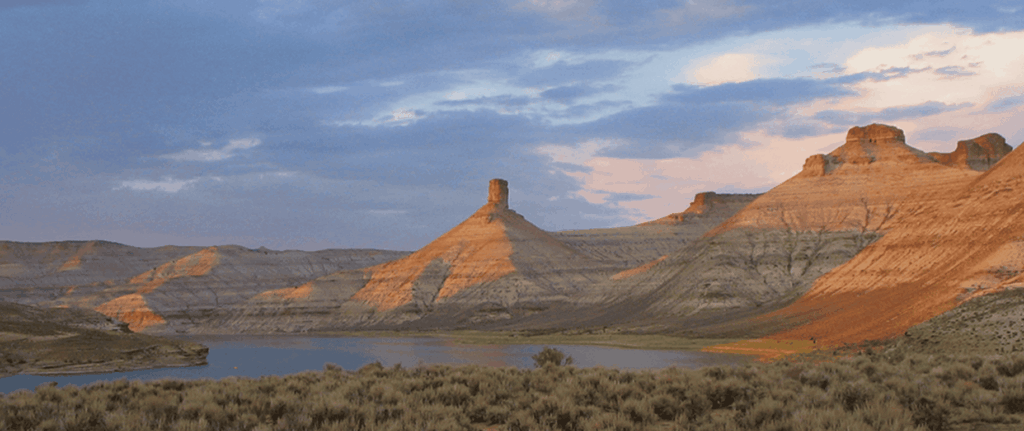 Desert landscape with layered mesas, a central butte, and a lake in the foreground under a partly cloudy sky at sunset.