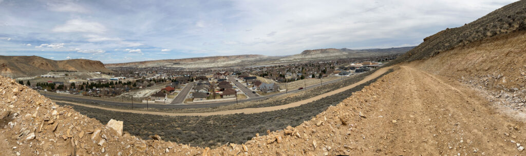 Panoramic view of a small town with houses and roads, seen from a rocky hill under a partly cloudy sky.
