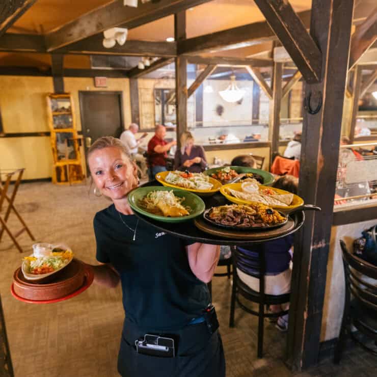 A server smiles while carrying several plates of food in a busy restaurant with wood accents and diners seated at tables in the background.