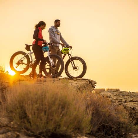 Two people with mountain bikes stand on a rocky cliff at sunset, overlooking the landscape.
