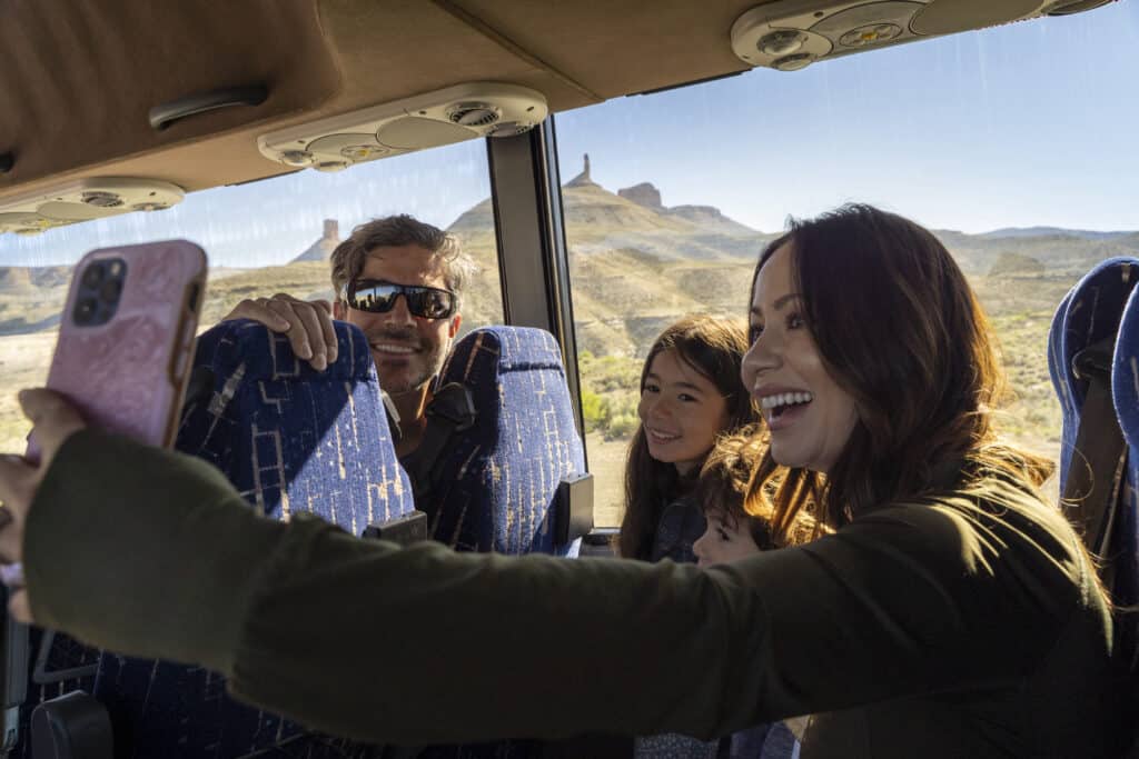 Four people sit together on a bus, smiling and taking a selfie with a smartphone. Sunlight streams in through the window, and desert scenery is visible outside.