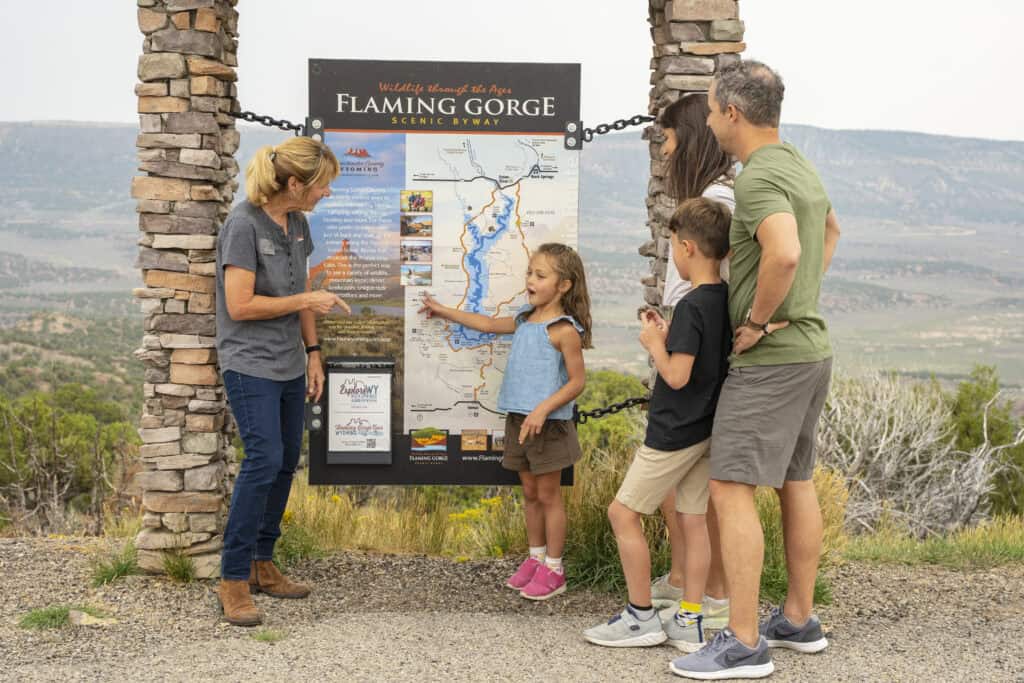 A woman points to a map of Flaming Gorge as a family of four listens and looks at the sign, with mountains and landscape in the background.