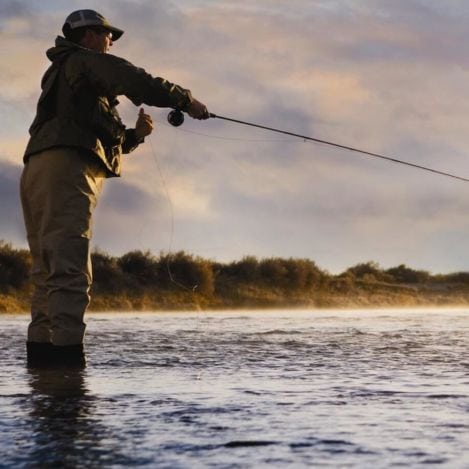 Person standing in shallow water fly fishing at sunrise or sunset, wearing outdoor gear and a cap.