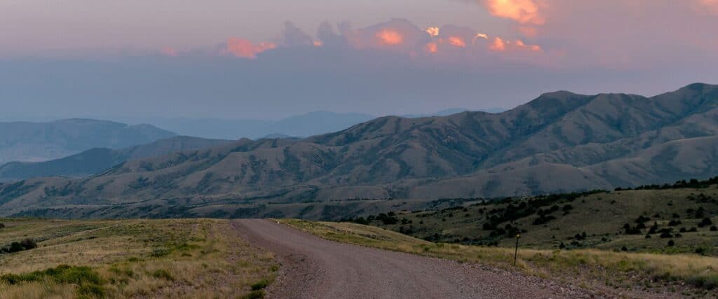 A gravel road curves through grassy hills with mountains in the background under a cloudy sky at sunset.