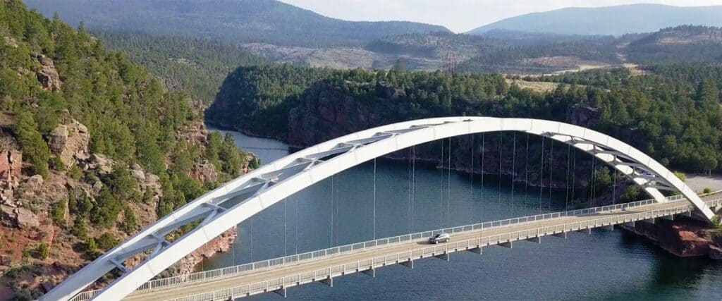 A white arch bridge spans a river surrounded by forested hills and mountains under a partly cloudy sky.