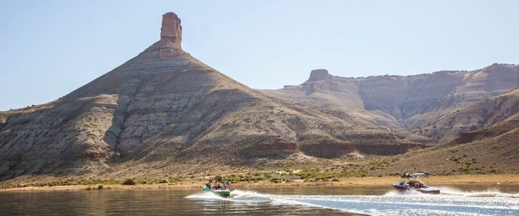 Two motorboats speed along a calm lake near a barren, rocky hill with a tall, narrow rock formation at its peak under a clear sky.