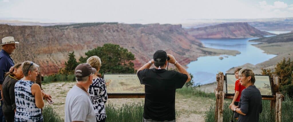 A group of people stand at an overlook reading informational signs, with a canyon and river visible in the background.