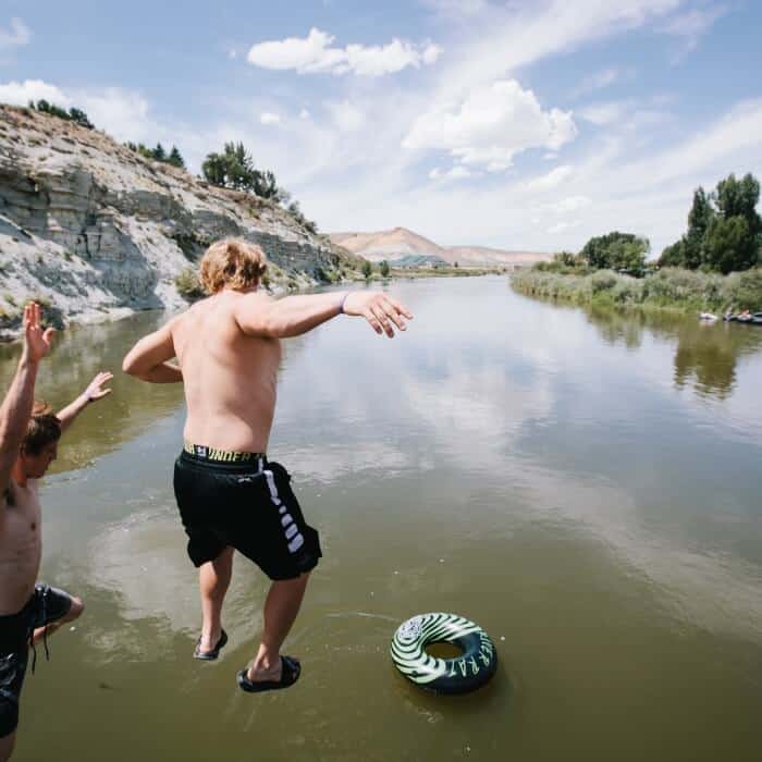 Two people in swimwear jump into a river from the shore, with a green inflatable ring floating in the water below them. Rocky banks and trees line the background under a partly cloudy sky.