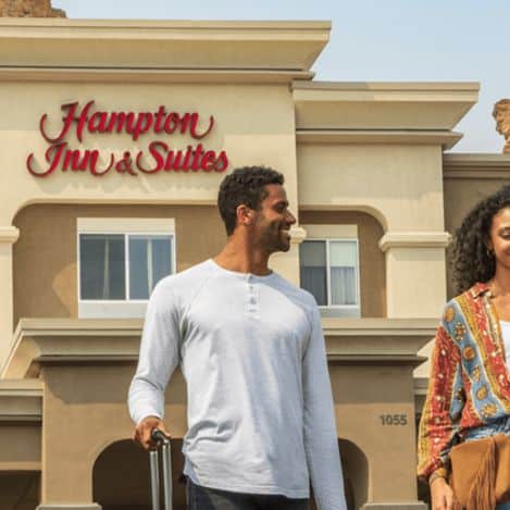 A man and woman stand outside a Hampton Inn & Suites hotel, with the building and hotel sign visible in the background. The man is holding a suitcase.