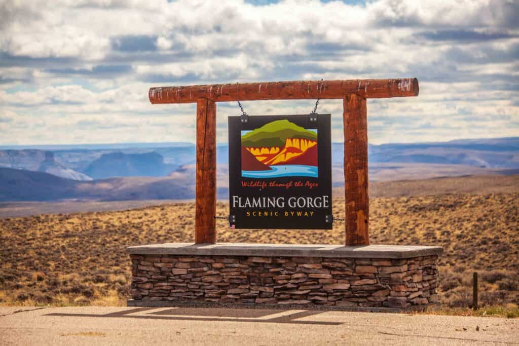 A wooden sign for Flaming Gorge Scenic Byway stands in a dry, open landscape with rolling hills and cloudy sky in the background.