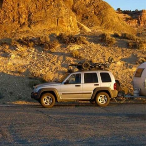 A silver SUV with a bicycle on its roof is parked next to a small trailer in a rocky, sunlit desert landscape.