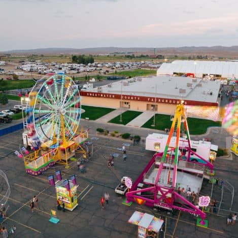 Aerial view of a fairground with a Ferris wheel, amusement rides, food stalls, and crowds outside the Sweetwater County Fairgrounds building.