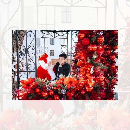 A young boy sits with Santa Claus in a display decorated with red tinsel, ornaments, and flowers.