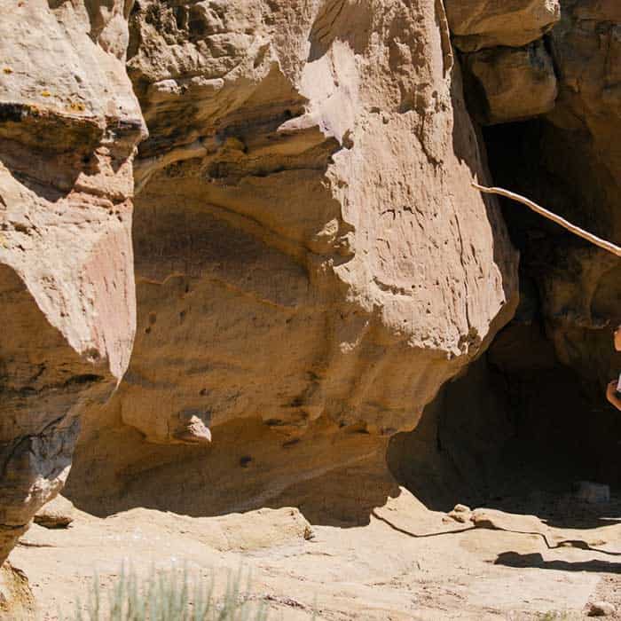 A person holding a stick stands near large, sunlit rock formations with deep shadows and textured surfaces.