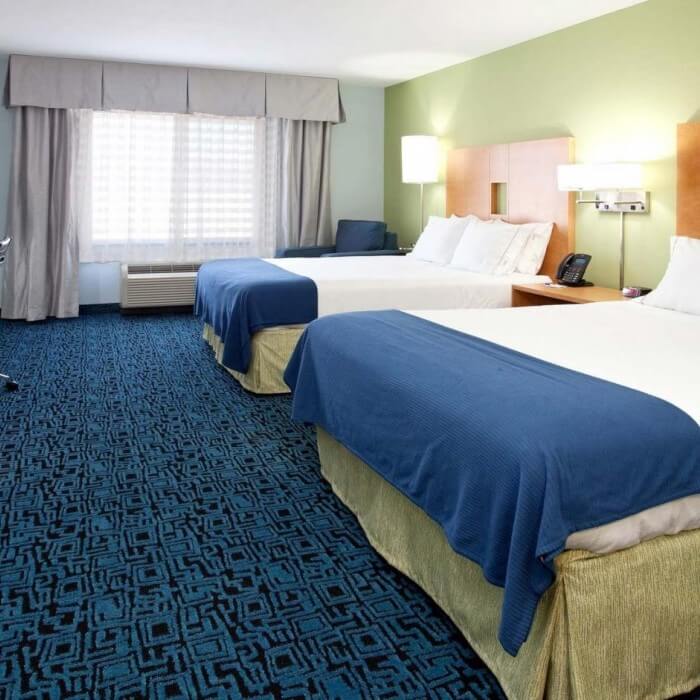 Hotel room with two neatly made double beds, blue bedspreads, patterned blue carpet, a window with drapes, and a small desk with a chair in the corner.