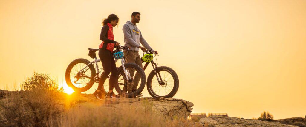Two people with mountain bikes stand on a rocky hilltop at sunset, looking into the distance.