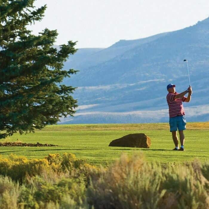 A man in a red striped shirt and blue hat swings a golf club on a grassy golf course with mountains in the background.