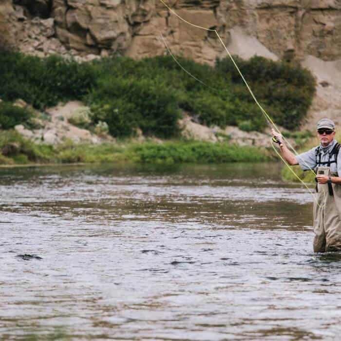 Person wearing fishing gear stands in a shallow river, casting a fishing line with a fly rod. Rocky terrain and greenery are visible in the background.